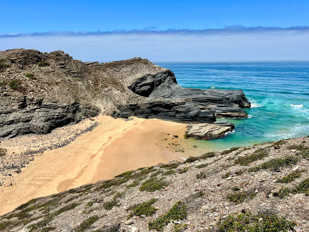 A dark stoned cliff along a beach in contrast of the light blue color of the ocean